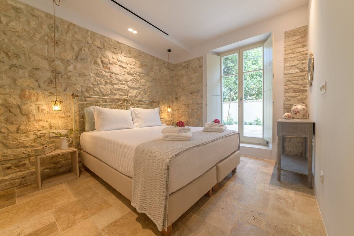 A bedroom with a stone wall, a large bed with white linens, a bedside table with a lamp, and a window with a view of greenery.