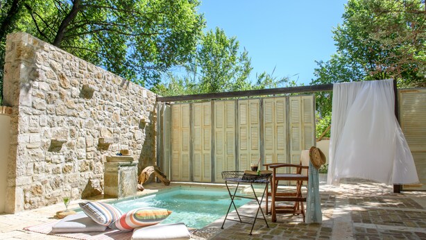 A stone wall, a small pool, and a table with chairs in a sunny outdoor area. - Carpofoli Corfu (Corfu)