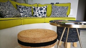 Une pièce avec un canapé jaune, un tabouret en osier et une petite table ronde à structure métallique.