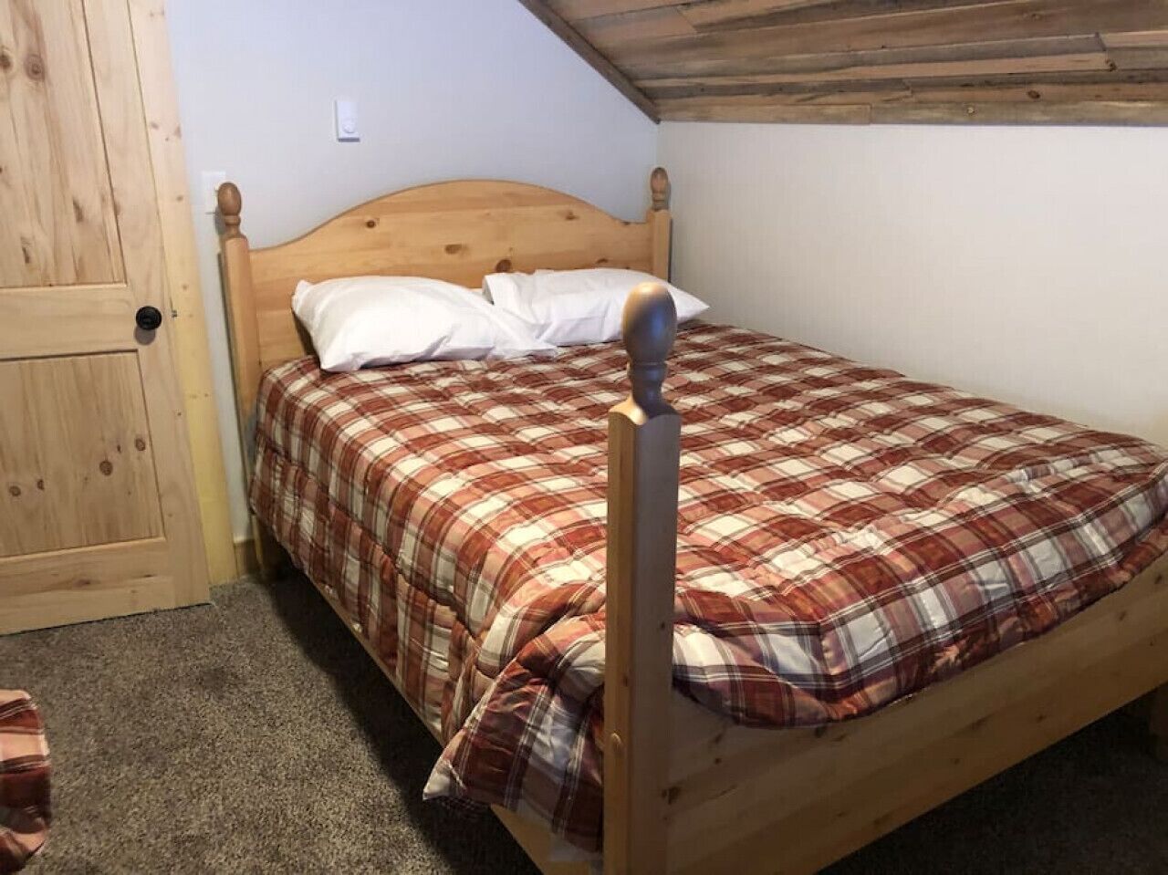 A single wooden bed with a plaid bedspread and white pillows in a room with a wooden door and a sloped ceiling.