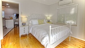 Une chambre avec un lit en métal blanc, deux tables de chevet avec des lampes, un ventilateur de plafond blanc et une fenêtre avec des stores.