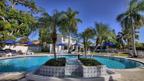 A swimming pool with tiled borders, surrounded by lounge chairs and blue umbrellas, with palm trees and a building in the background. - Pool view.  Upstairs unit & workspace (St Pete Beach)