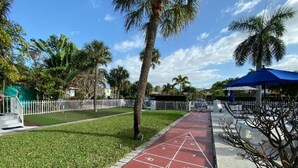 A sunny outdoor area with a red pavement for shuffleboard, surrounded by palm trees and a white fence.
