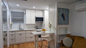 Een moderne keuken met witte kasten, een fornuis en een spoelbak. Er is een eethoek met een witte tafel en stoelen, en een bruine bank in de hoek.