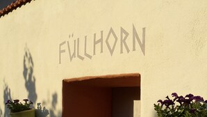 A building facade with the name "FULLHORN" in large letters. There are two hanging planters with plants on either side of a door.