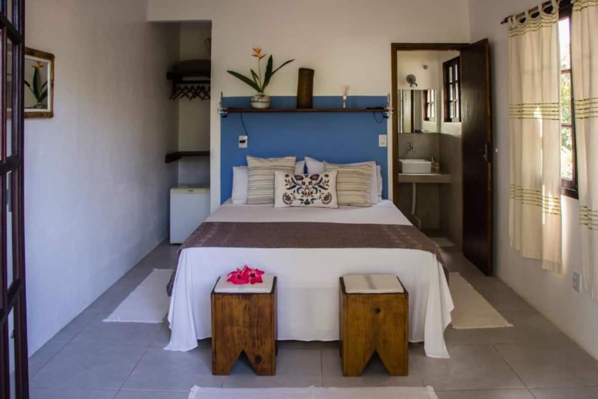 A bedroom with a bed, bedside tables, a nightstand, a shelf with decorative items, a window with curtains, and a door.