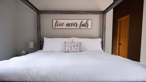 Una cama bien hecha con ropa blanca y dos almohadas, una con el texto "HAPPY PLACE". Se ve un cabecero de madera y un cartel en la pared que dice "love never fails".