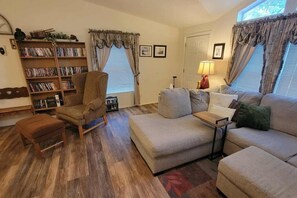 Ein Wohnzimmer mit Sofa, Sessel, Hocker, Bücherregal und Lampe.