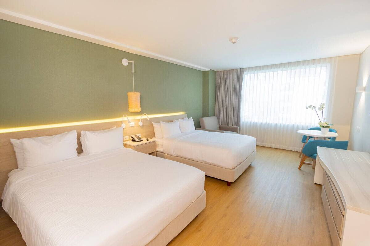 Une chambre d’hôtel avec deux lits, un bureau, une chaise, une lampe et une fenêtre avec des rideaux.