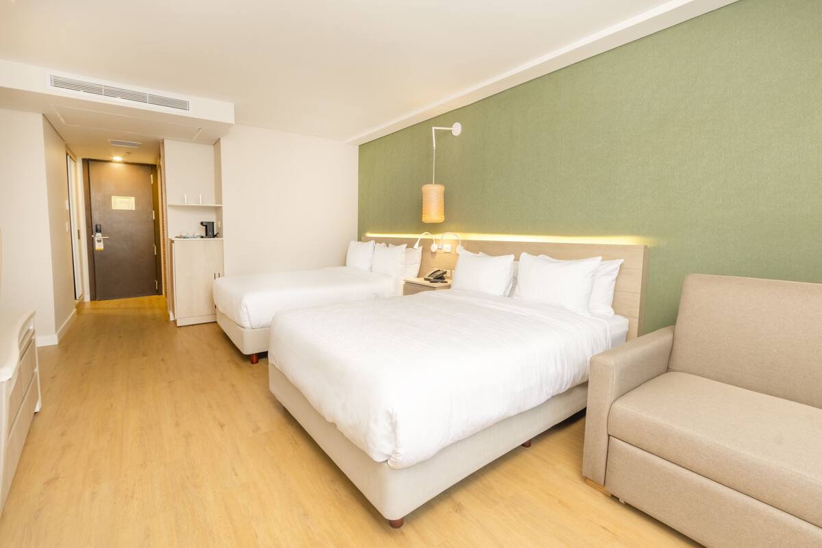 Une chambre d’hôtel avec deux lits, un canapé, une table de chevet avec une lampe et un mur d’accent vert.