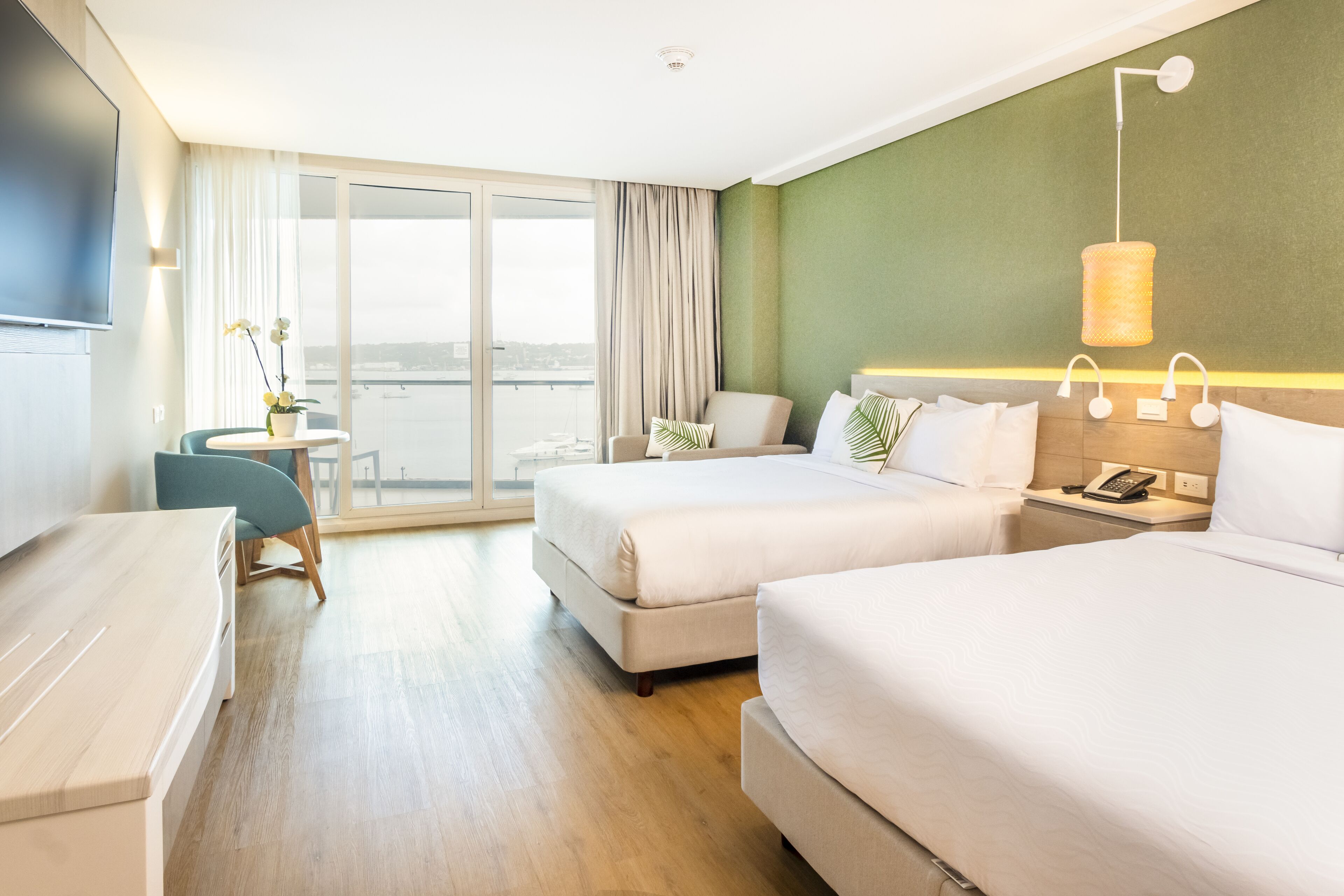 Habitación de hotel con dos camas, un escritorio, una silla, una mesita con un florero y vistas de la ciudad a través de amplios ventanales.