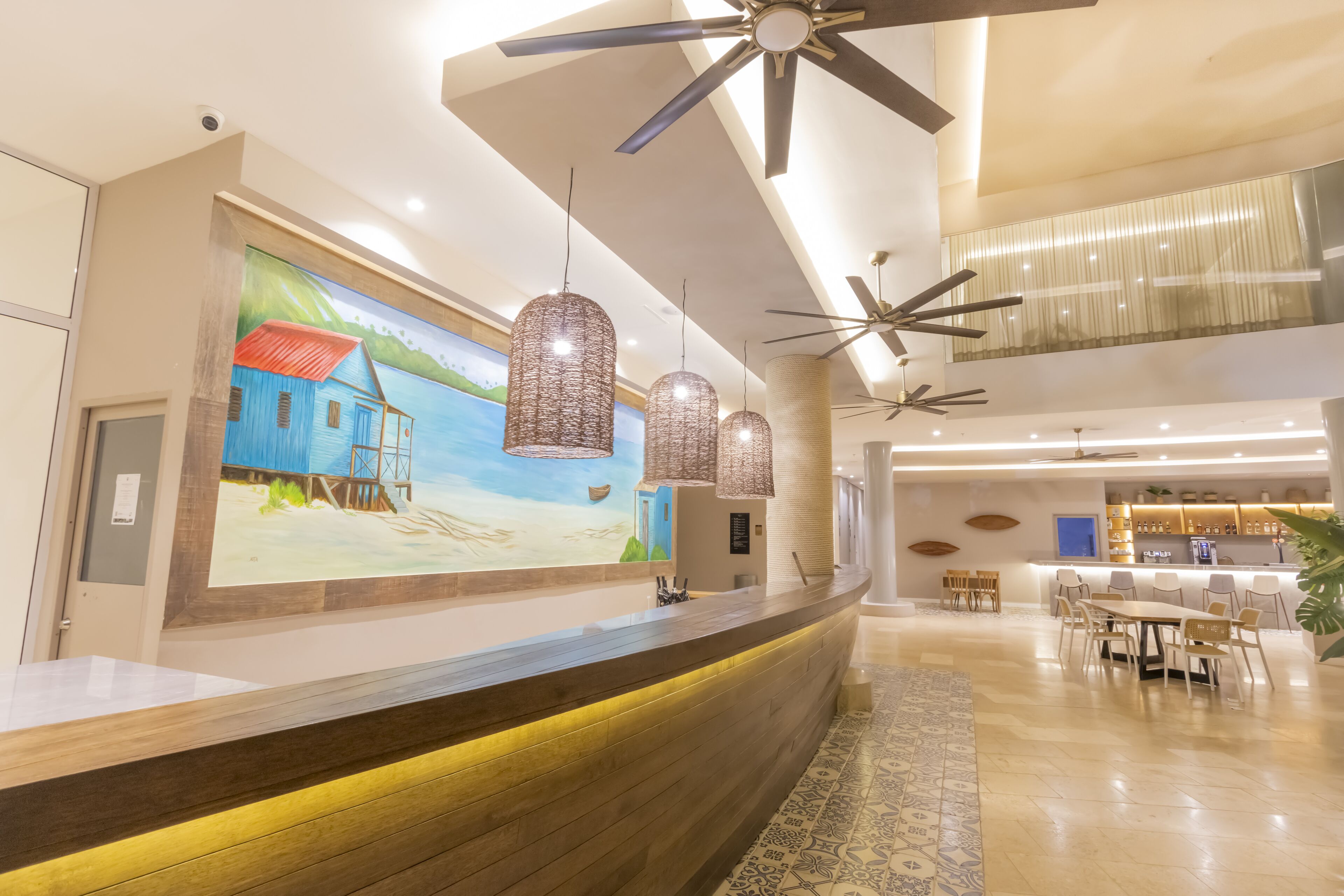 A hotel lobby with a reception desk, pendant lights, ceiling fans, a mural, and a dining area.