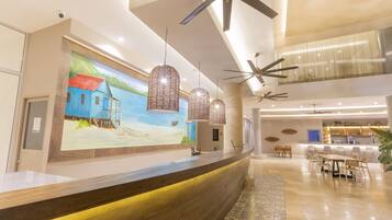 A hotel lobby with a reception desk, pendant lights, ceiling fans, a mural, and a dining area.