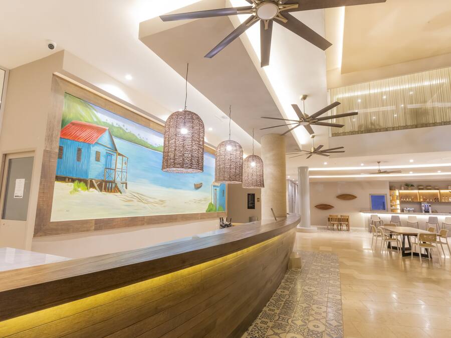 A hotel lobby with a reception desk, pendant lights, ceiling fans, a mural, and a dining area.