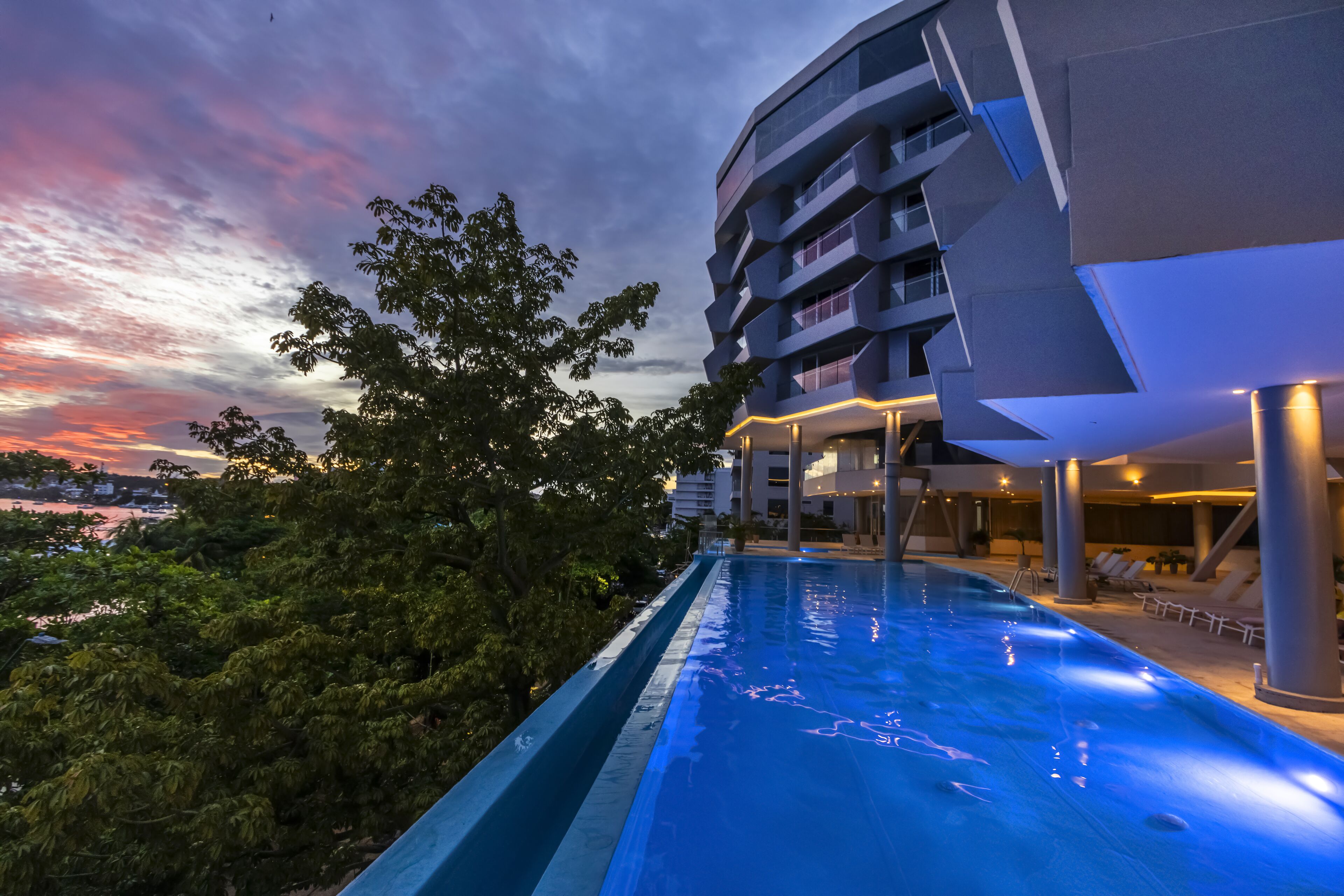 A modern hotel with a swimming pool, surrounded by lush greenery and a view of the cityscape at dusk.