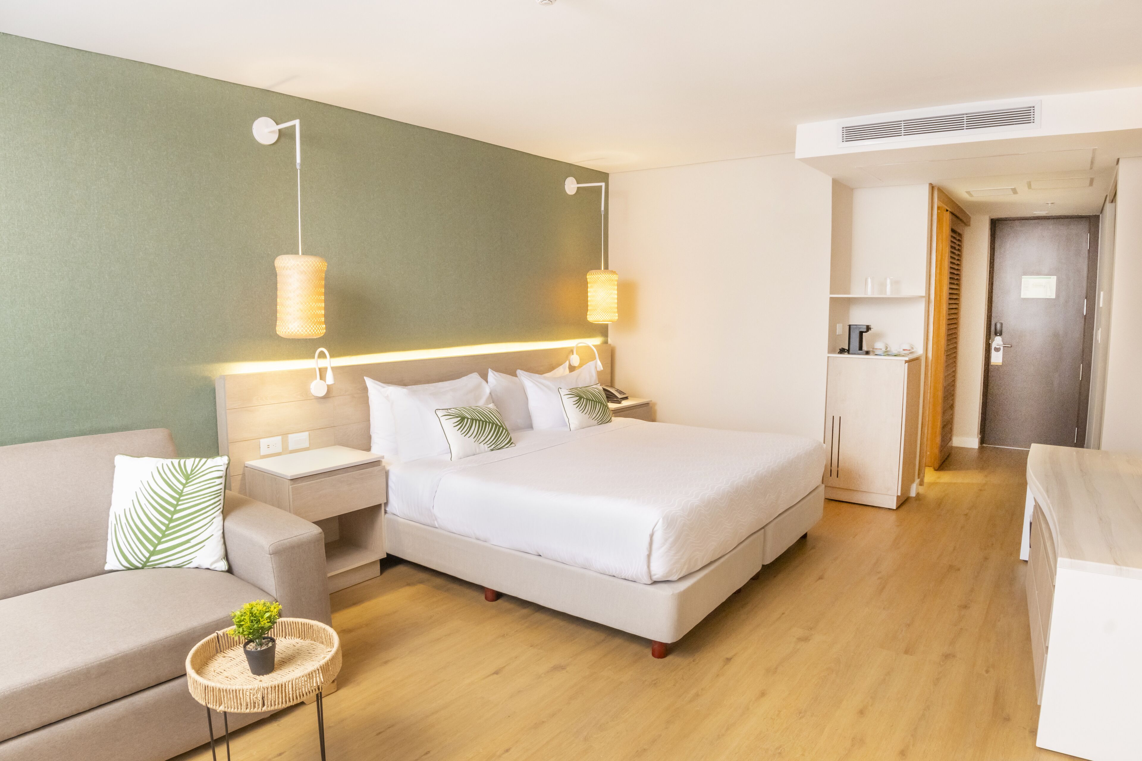 A modern hotel room with a bed, bedside tables, a sofa, a small table with a plant, and a wardrobe.