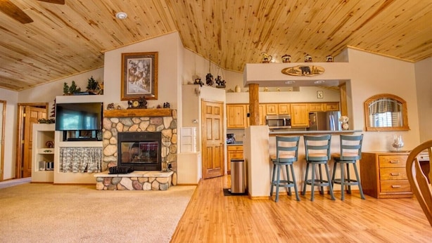 A cozy living room with a stone fireplace, wooden ceiling, and a kitchen area with bar stools.
