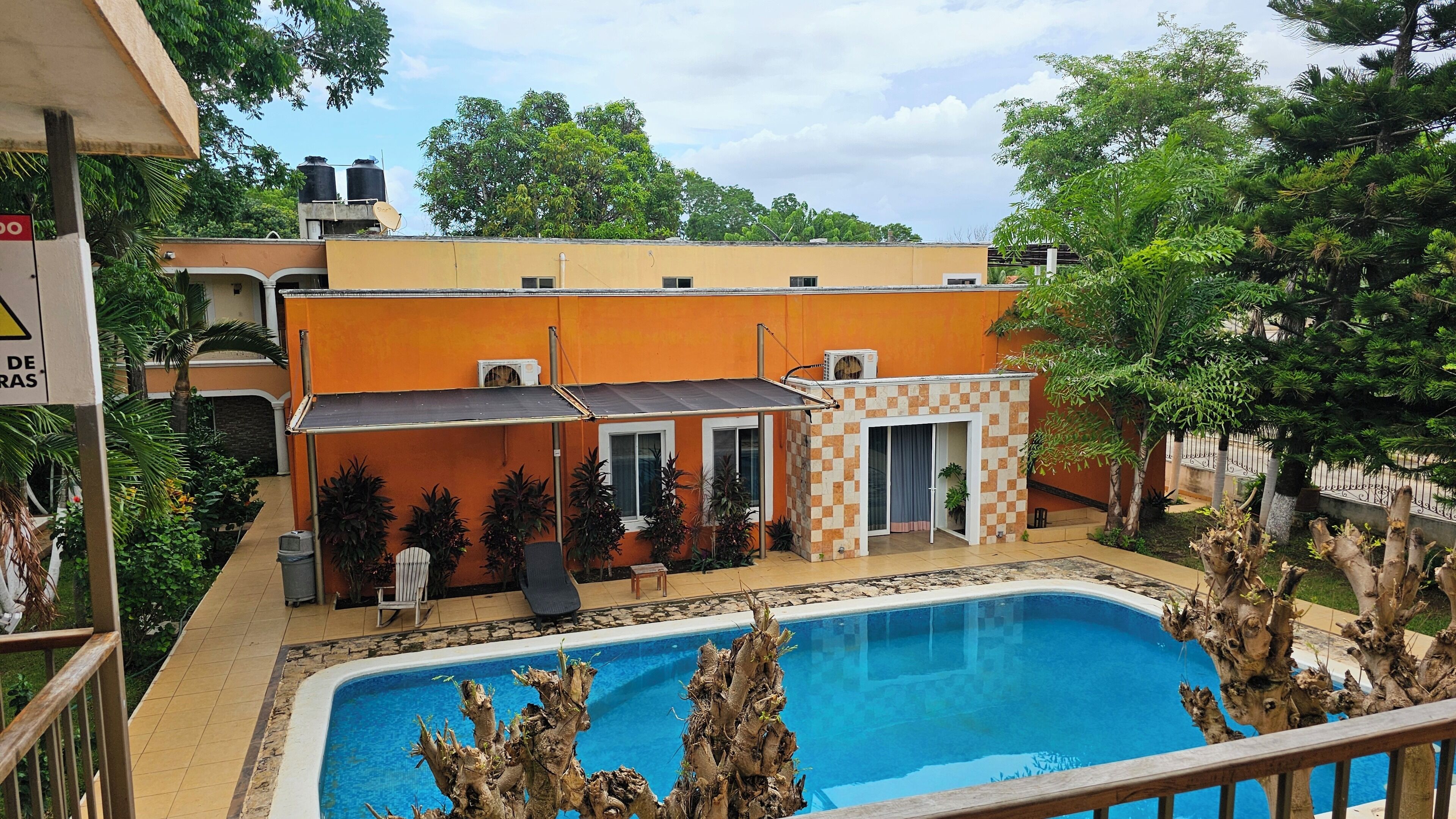 En swimmingpool med flisebelagt terrasse, omgivet af et hegn med en skakternet bygning og et overdækket siddeområde.