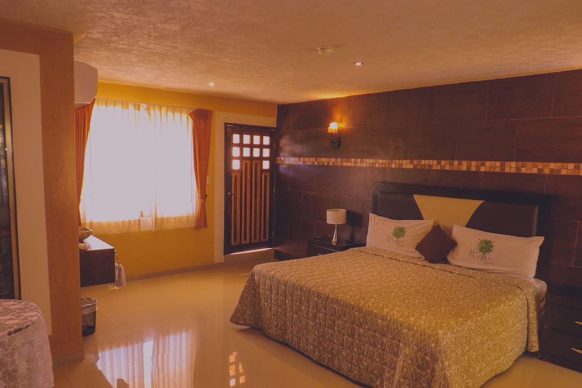 Un dormitorio con una cama grande, una mesita de noche con lámpara, una ventana con cortinas y suelo de baldosas.