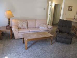 Un salon avec un canapé beige, une table basse en bois, un fauteuil foncé et une lampe sur une table d’appoint.