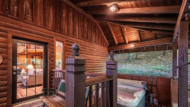 Una cabaña de troncos con porche de madera, bañera de hidromasaje y vistas al bosque.