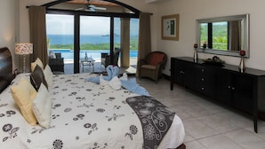 A bedroom with a large bed, a ceiling fan, a view of the ocean, a dining area with a table and chairs, and a black console table with a mirror and candles. - Casa Antonelli (Sardinal)