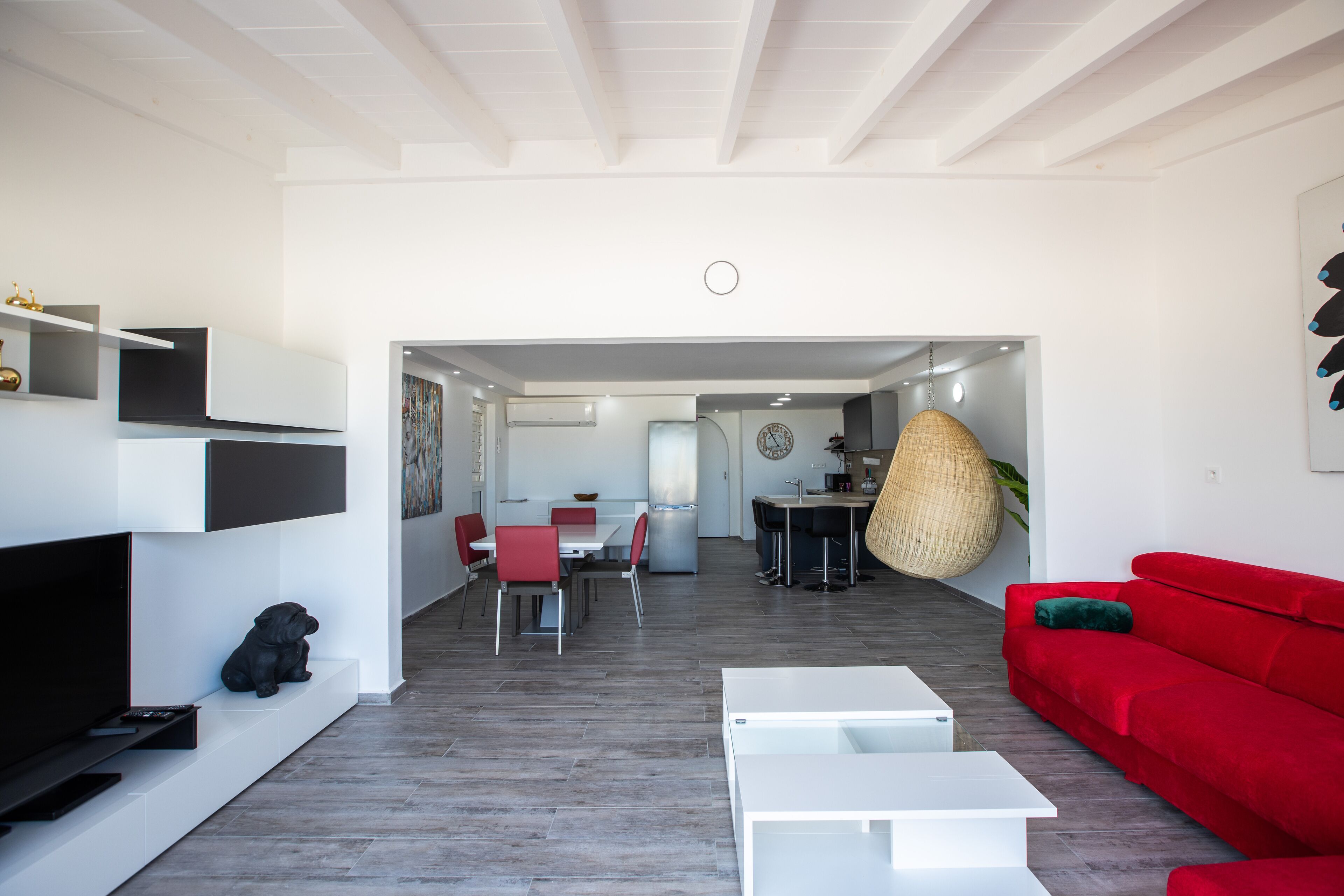 A modern living room with a red sofa, white coffee table, and a TV on a white stand. There is a dining area with red chairs and a table, and a kitchen visible in the background.