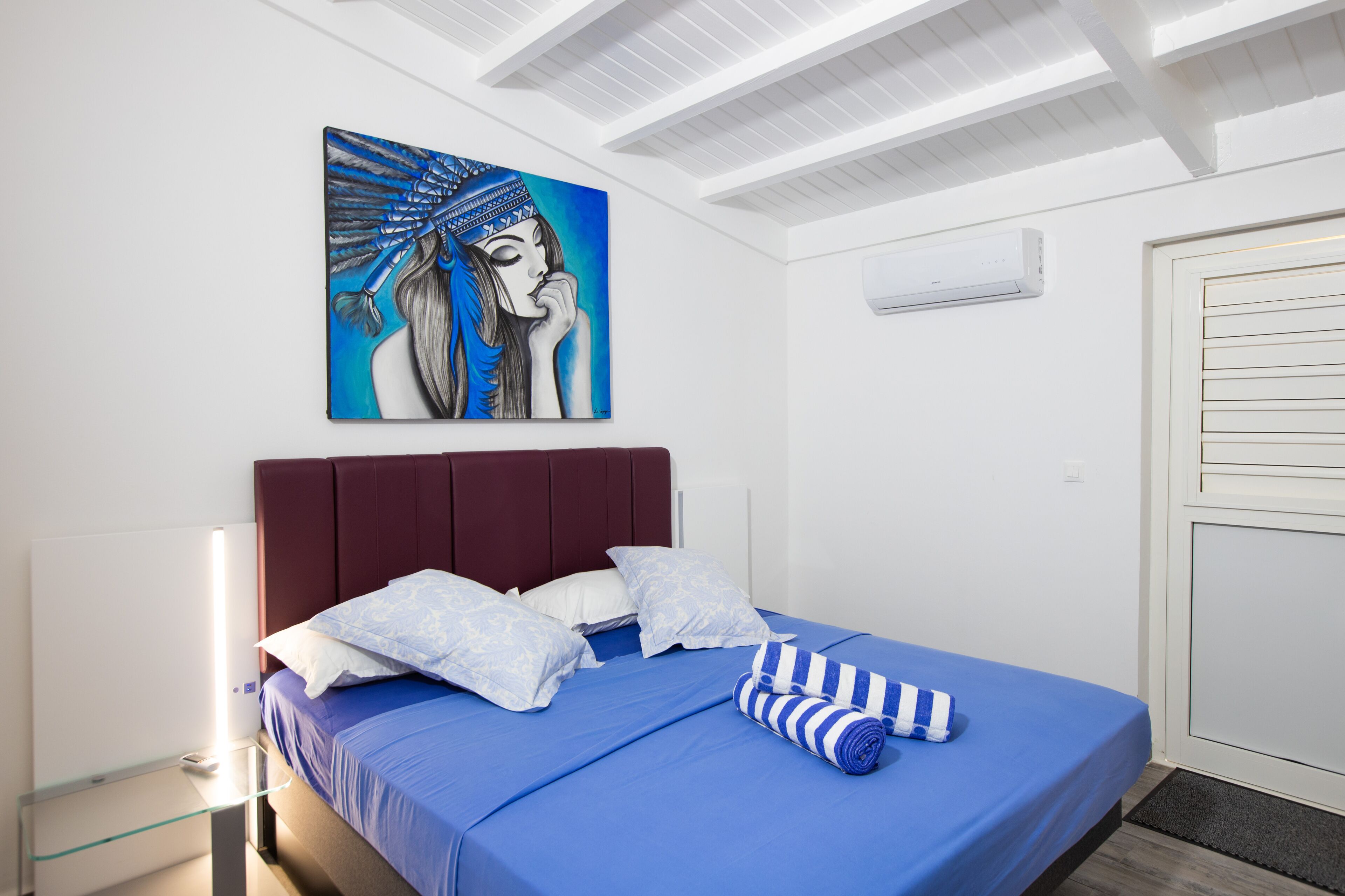 A hotel room with a bed, pillows, a painting, an air conditioning unit, and a door.