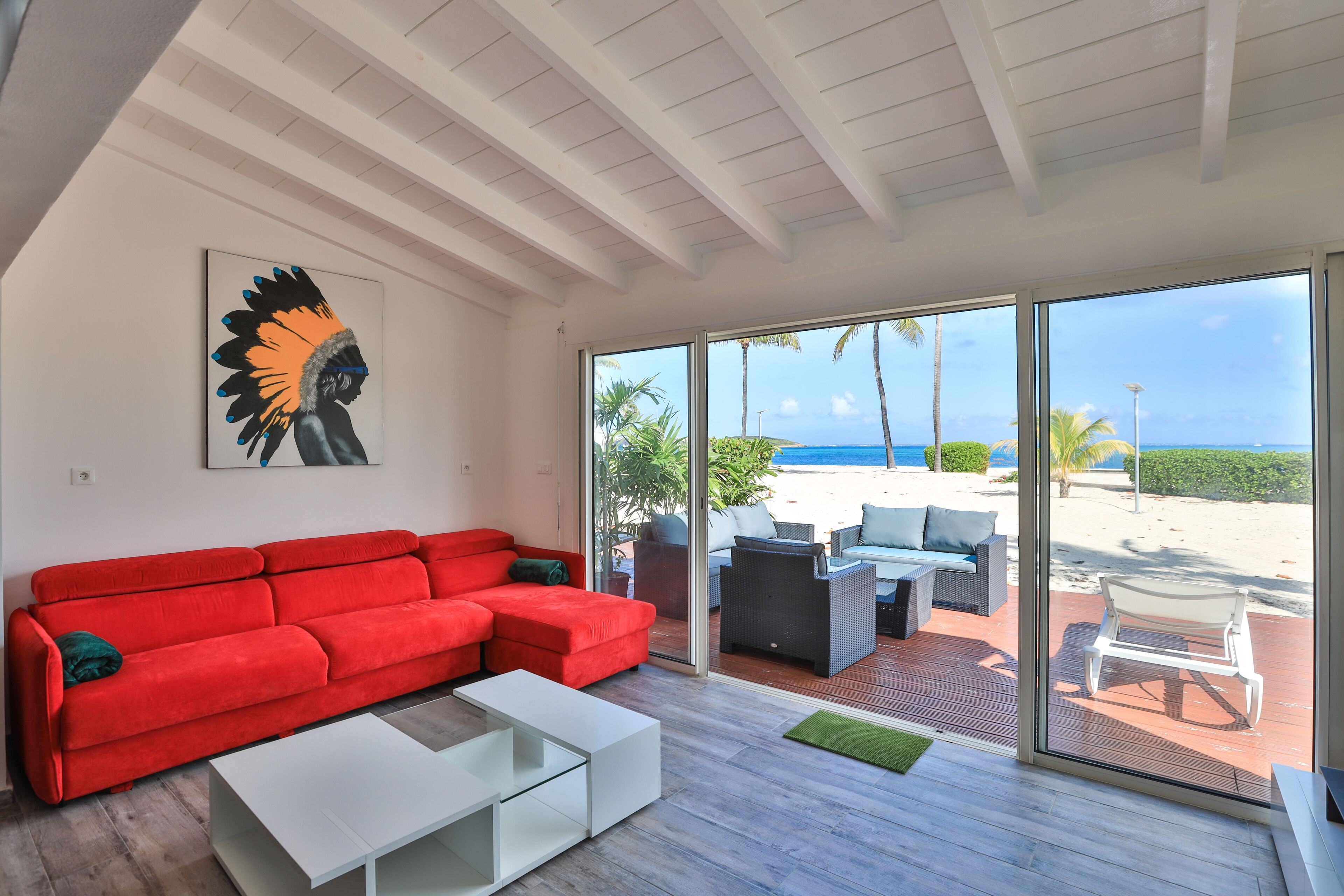 A modern living room with a red sofa, white coffee table, and a glass door leading to a deck with outdoor furniture and a view of the beach.
