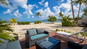 A beachside patio with wicker furniture, a white lounge chair, and a view of the ocean and mountains. - ZEN Apartment T2 "Waterfront" Baie Nettle (BAIE NETTLE)