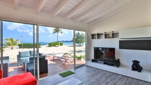 A modern living room with a large glass door leading to a beach view, a flat-screen TV, and a red sofa. - ZEN Apartment T2 "Waterfront" Baie Nettle (BAIE NETTLE)