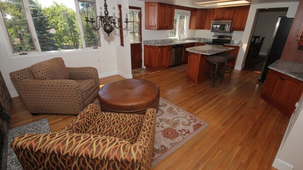 A spacious living room with a patterned sofa and ottoman, a kitchen with wooden cabinets and modern appliances, and large windows offering a view of the outdoors.