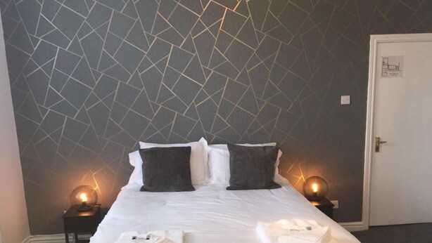 A hotel room with a geometric-patterned wall, a neatly made bed with white linens, two bedside tables with lamps, and a door. - The Waverley Hotel (Whitehaven)