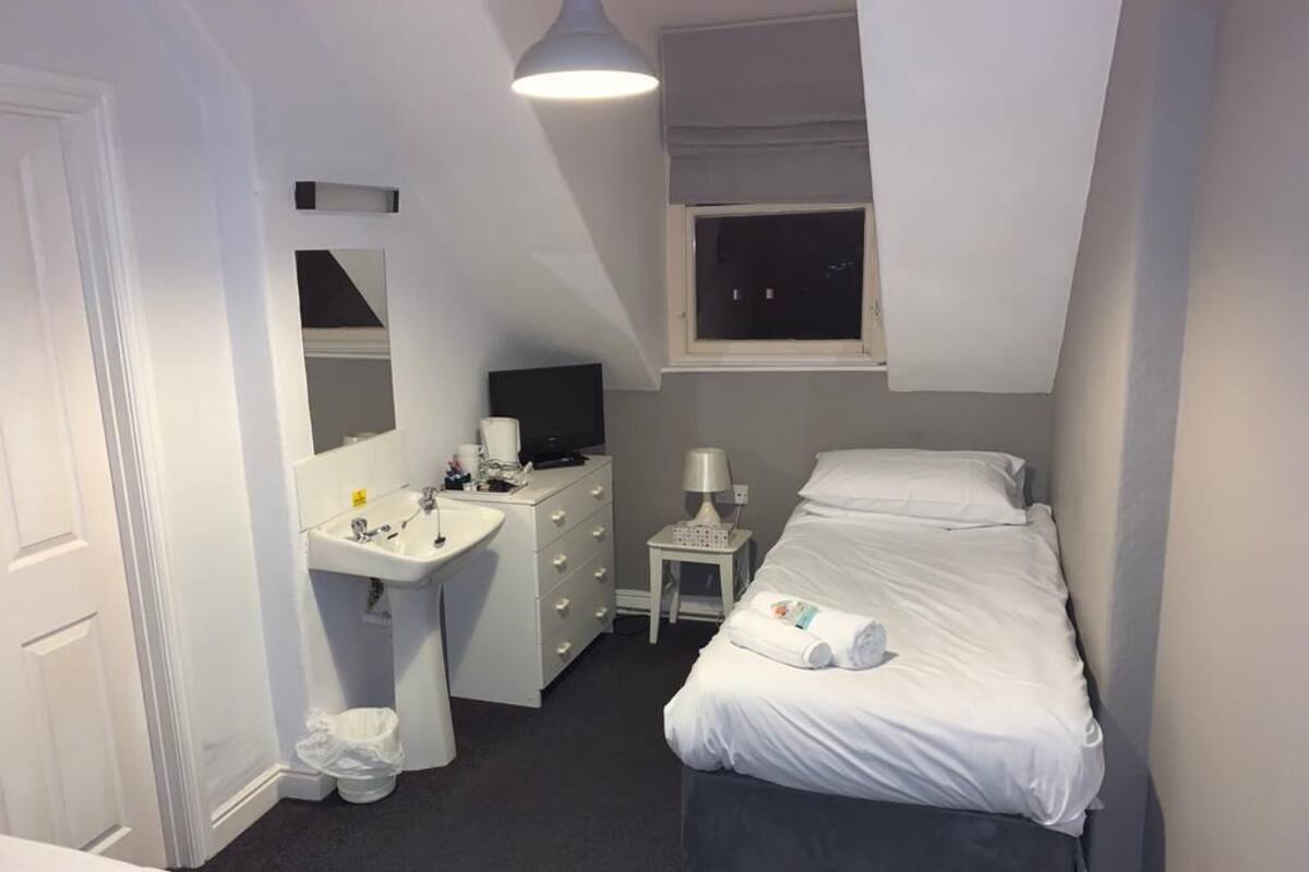 A small, well-lit bedroom with a bed, bedside table, and a desk with a mirror and sink.