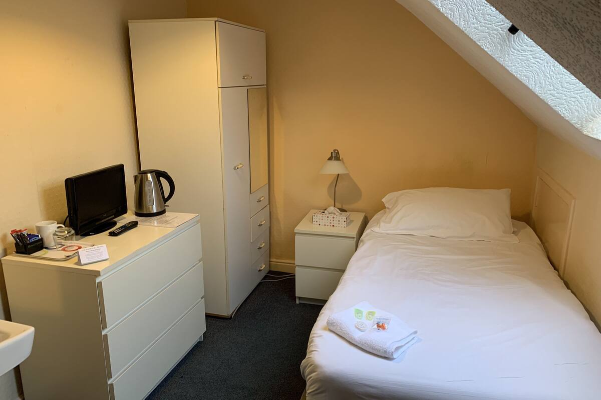 A small, neatly arranged bedroom with a bed, a desk with a computer, a kettle, and a nightstand with a lamp.