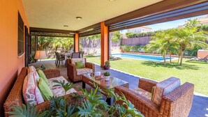 Un patio cubierto con mobiliario de mimbre, una piscina y vistas a un jardín y zona junto a la piscina.