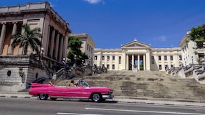 Une voiture décapotable rose est garée dans une rue, avec des bâtiments à l’architecture classique en arrière-plan.
