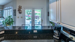 Una cocina con encimera de granito negro, cafetera y vista al jardín trasero por medio de puertas de vidrio.