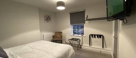 Un dormitorio con una cama, una silla, un radiador, una ventana y un televisor montado en la pared.