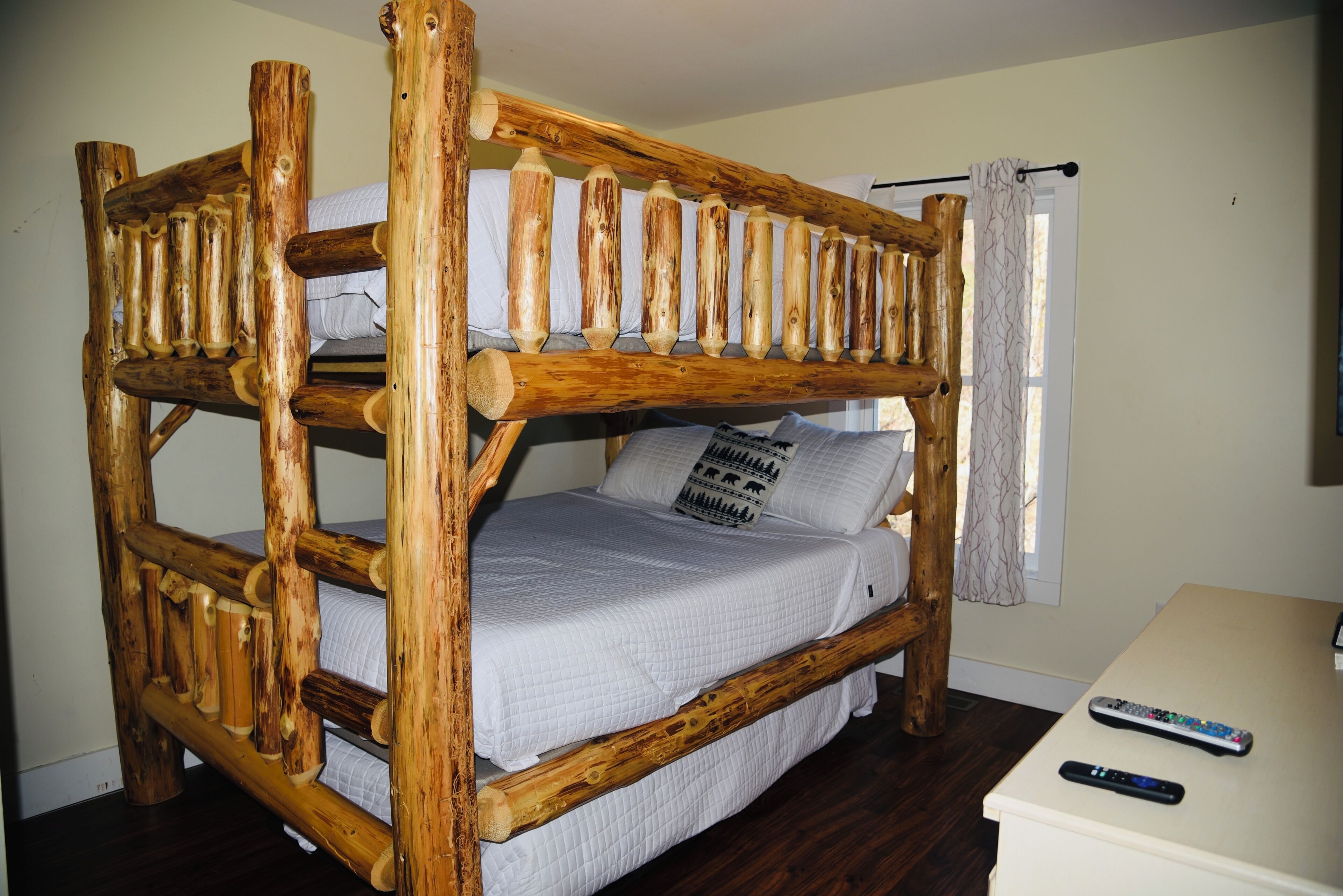 Una cama litera con estructura de madera y ropa de cama blanca. Hay un control remoto sobre una mesita de noche.