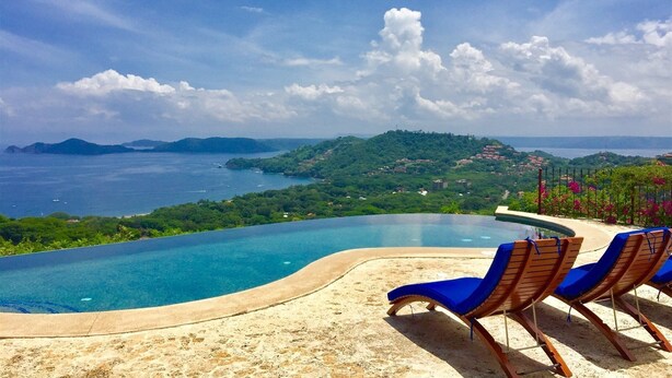Uma área à beira da piscina com espreguiçadeiras e vista para o mar e colinas.