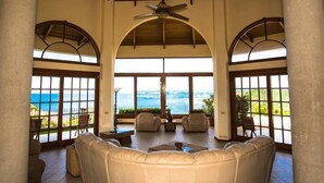 Uma sala de estar ampla com uma grande área de assentos acolchoados, um ventilador de teto e janelas amplas com vista para o mar.