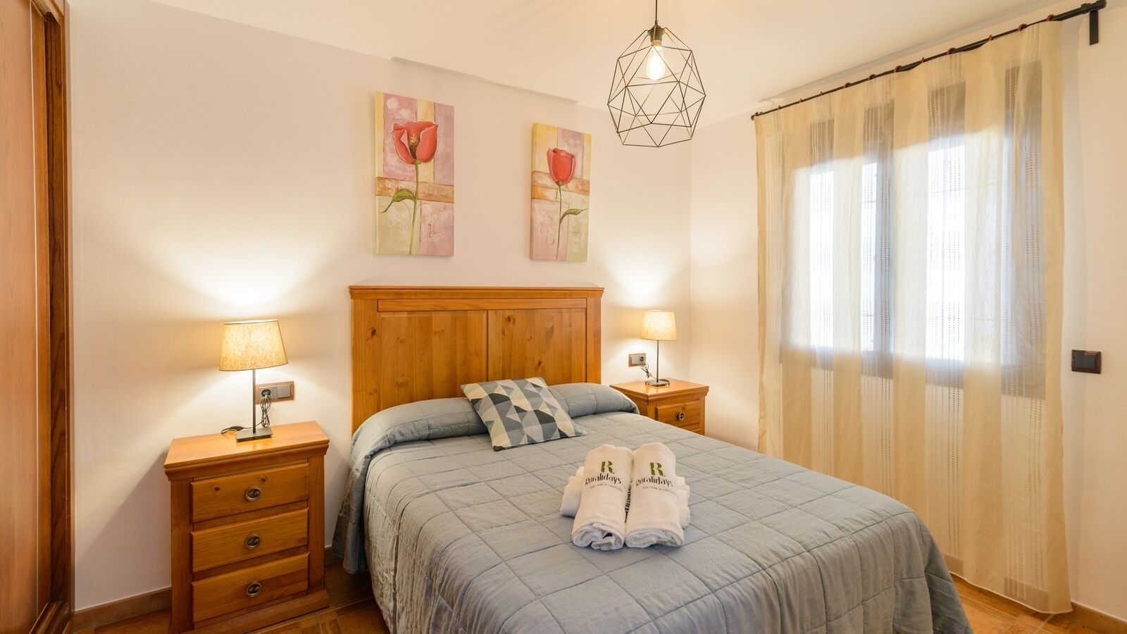 A bedroom with a wooden headboard, two bedside tables with lamps, a bed with a blue quilt, two paintings on the wall, and a window with sheer curtains.