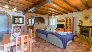 Un salon chaleureux avec un canapé, une table à manger et des chaises, une cheminée, et une cuisine en arrière-plan.