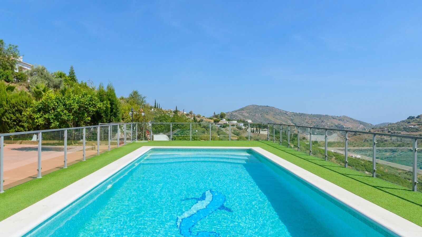 A rectangular swimming pool with a clear blue water feature, surrounded by a metal fence and green artificial grass.