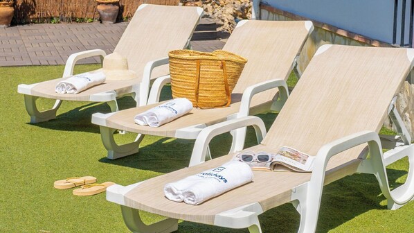 Three sun loungers with towels and a straw bag on a grassy area.