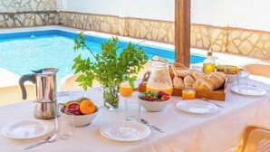 Une table de petit-déjeuner installée près d’une piscine, avec des corbeilles de fruits, une cafetière, du pain et du jus.
