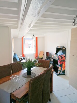 A dining area with a wooden table, wicker chairs, and a potted plant. There is a television and a small table with a decorative item in the background.