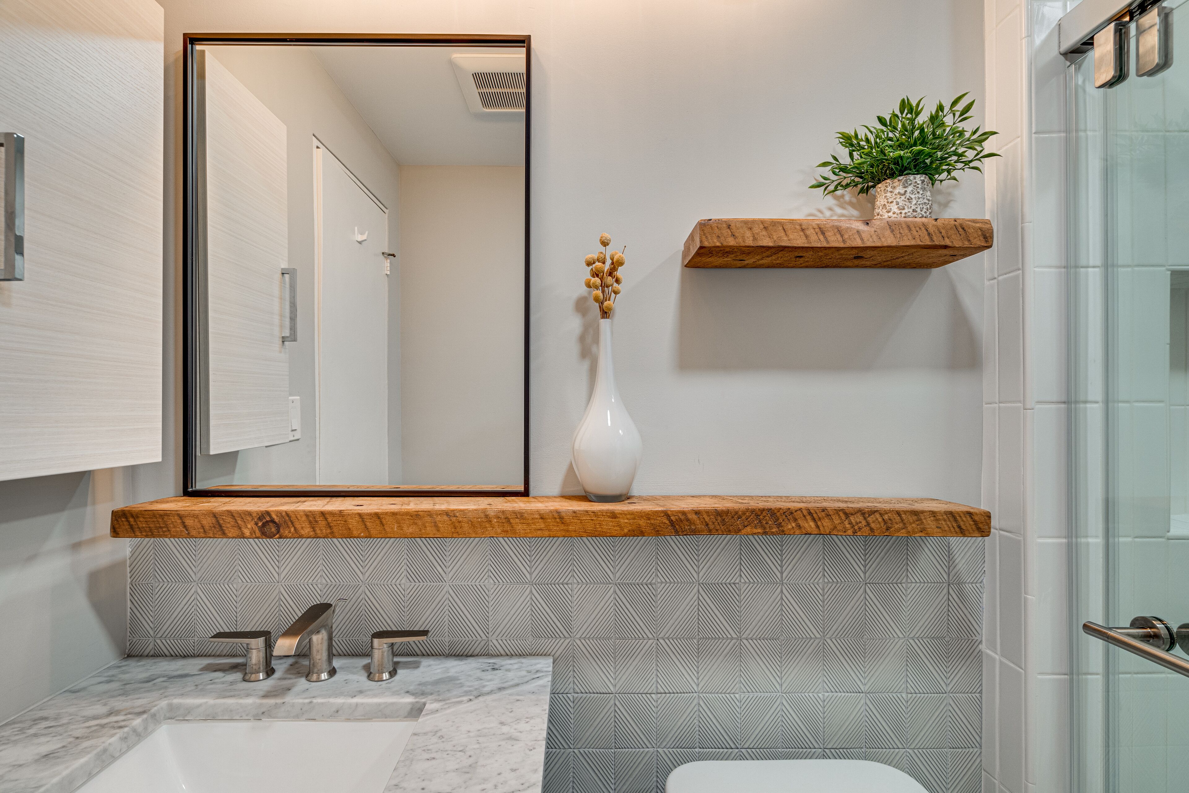 A bathroom with a wooden vanity, a white sink, a mirror, and a potted plant on a shelf.