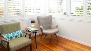 A room with a wooden floor, a green armchair with a patterned cushion, a grey upholstered chair, and a small table with a lamp and a book.